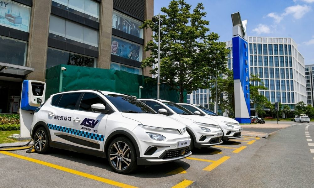 Dàn xe taxi ASV Airports màu xanh đang đỗ bên đường với cây xanh và bầu trời trong xanh