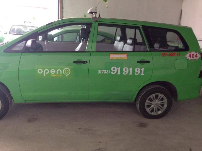 Hình ảnh xe taxi Open99 màu xanh lá cây đang di chuyển trên đường phố với các tòa nhà phía sau