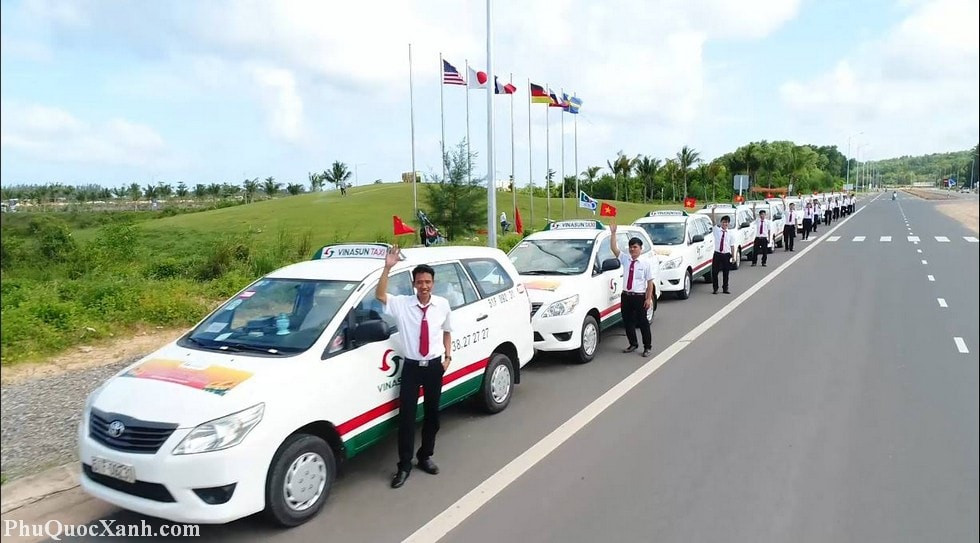 Một xe taxi màu đỏ đang chạy trên con đường ven biển đầy nắng, với cây cọ và các tòa nhà thấp tầng ở phía xa