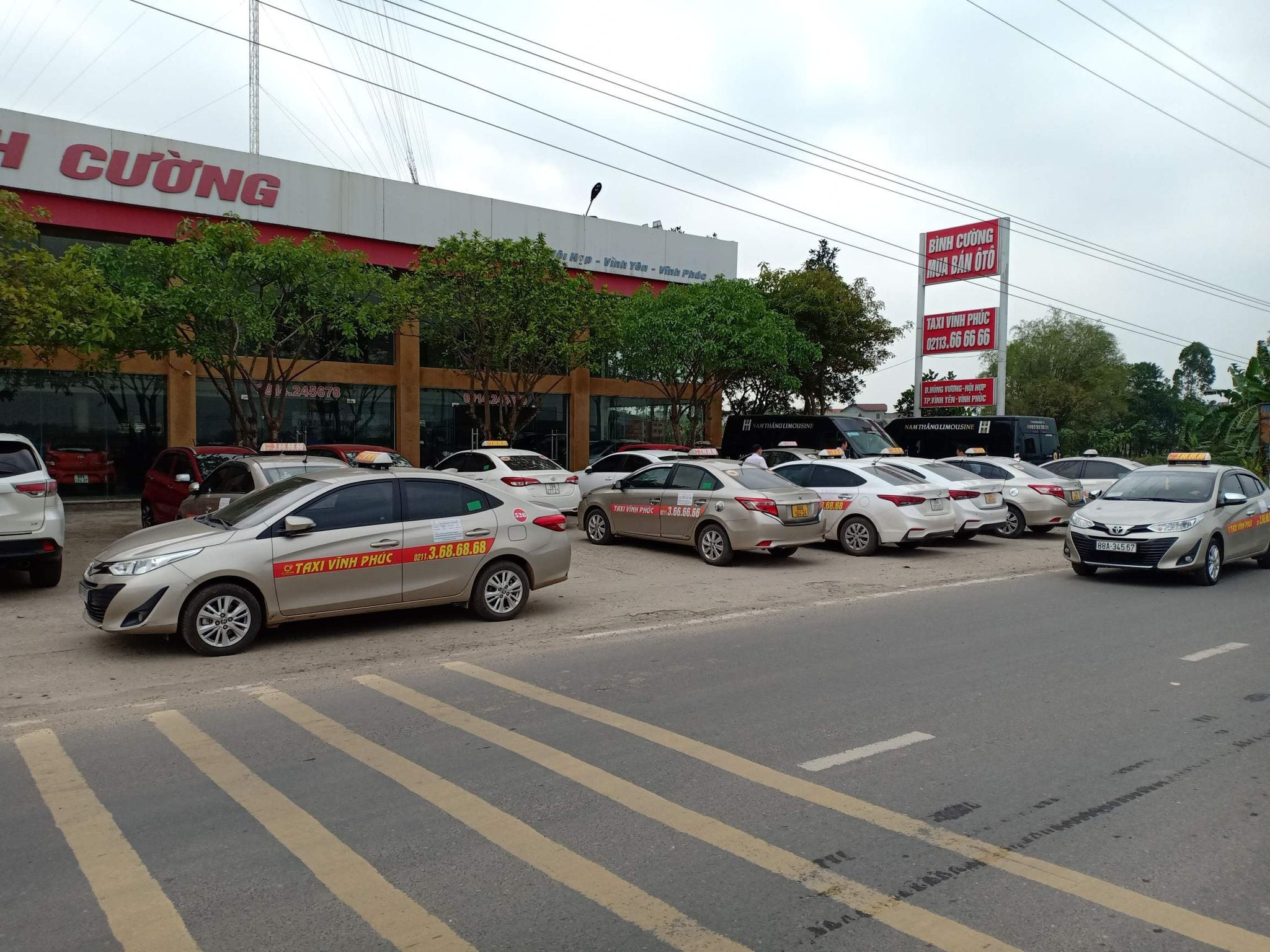 Xe taxi màu trắng đang đỗ trên đường, có chữ Taxi Yên Bái màu xanh và đỏ trên cửa xe. Phía sau là cảnh đồi núi và cây xanh, bầu trời trong xanh.